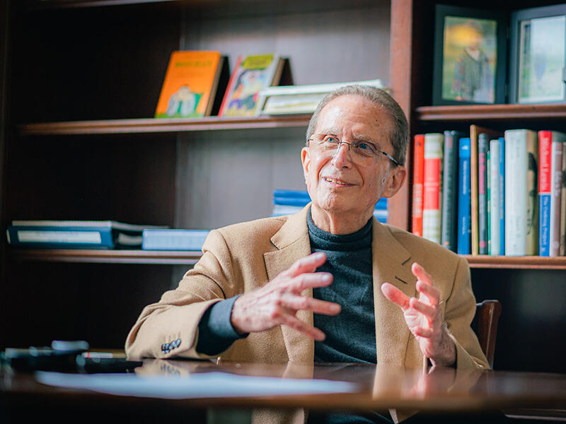 Michael Botchan gestures with his hands in front of a wooden bookshelf