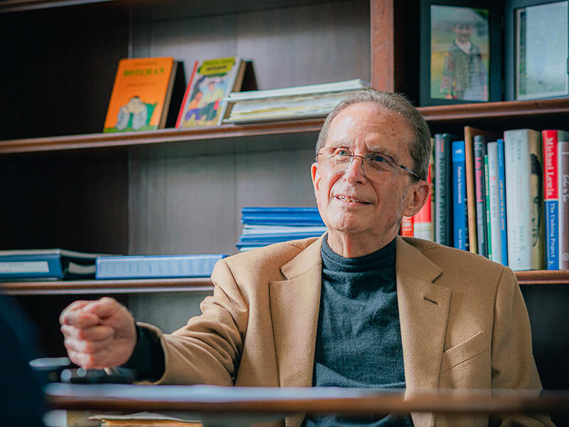 Michael Botchan rests his hand on a table in front of a wooden bookshelf