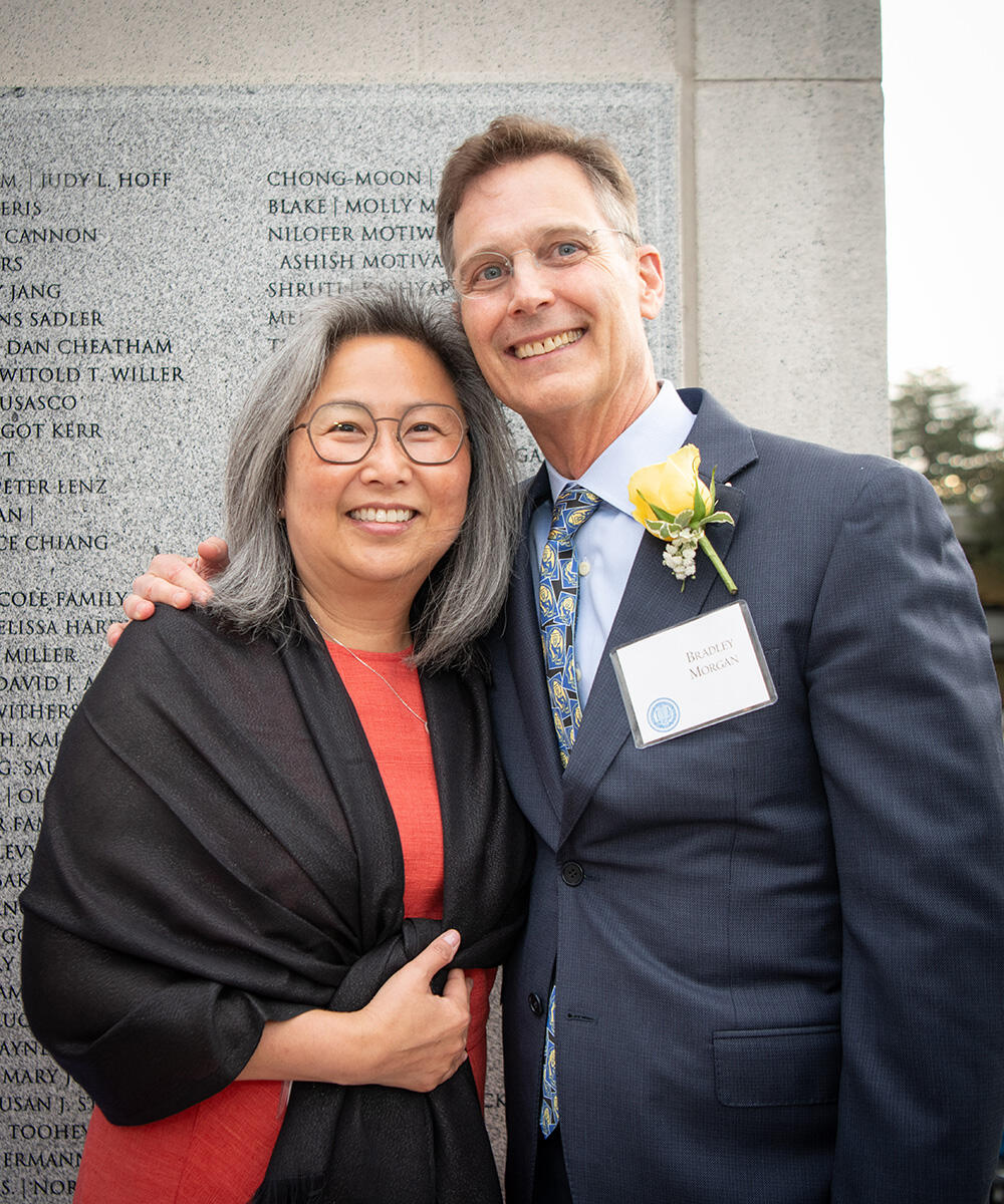 Julie and Brad, wearing formal attire, pose together in front of a granite wall with ethced names of other Builders of Berkeley