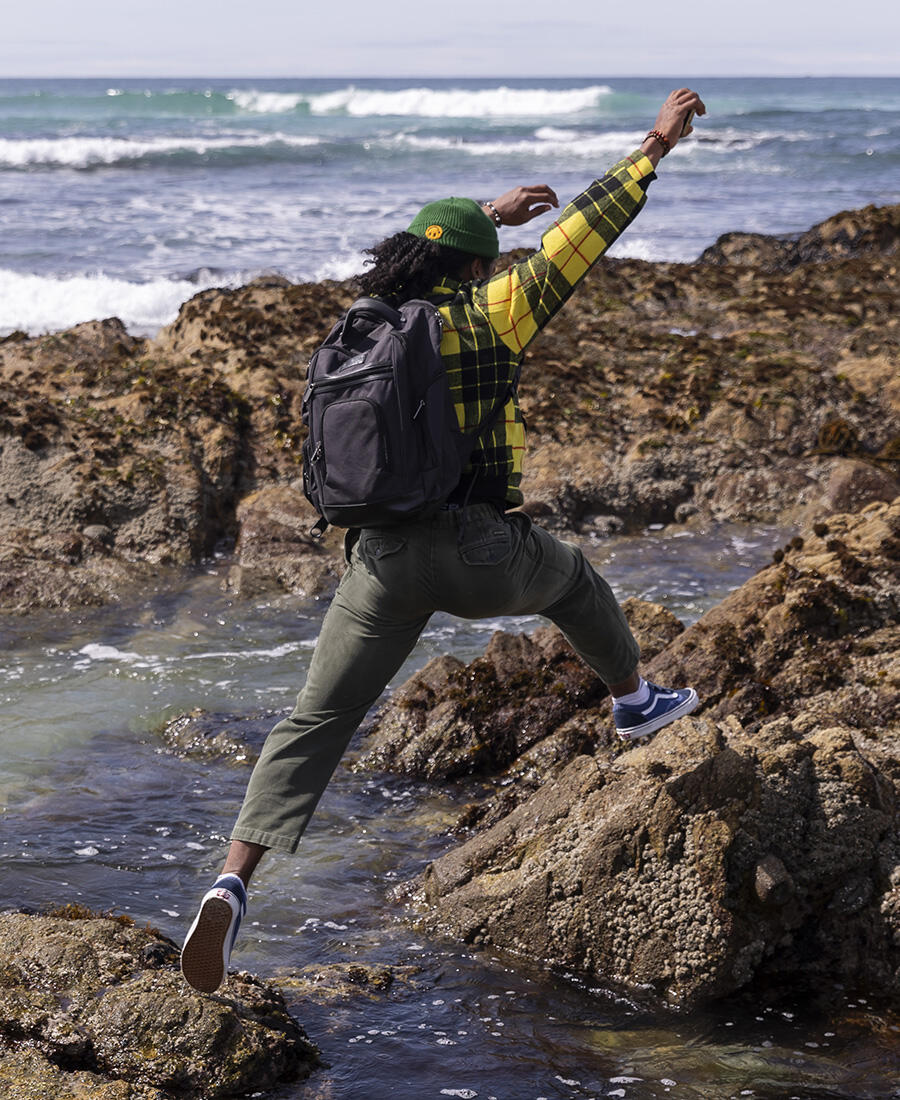 A student wearing a backpack jumps away from the camera to a rock at the beach.