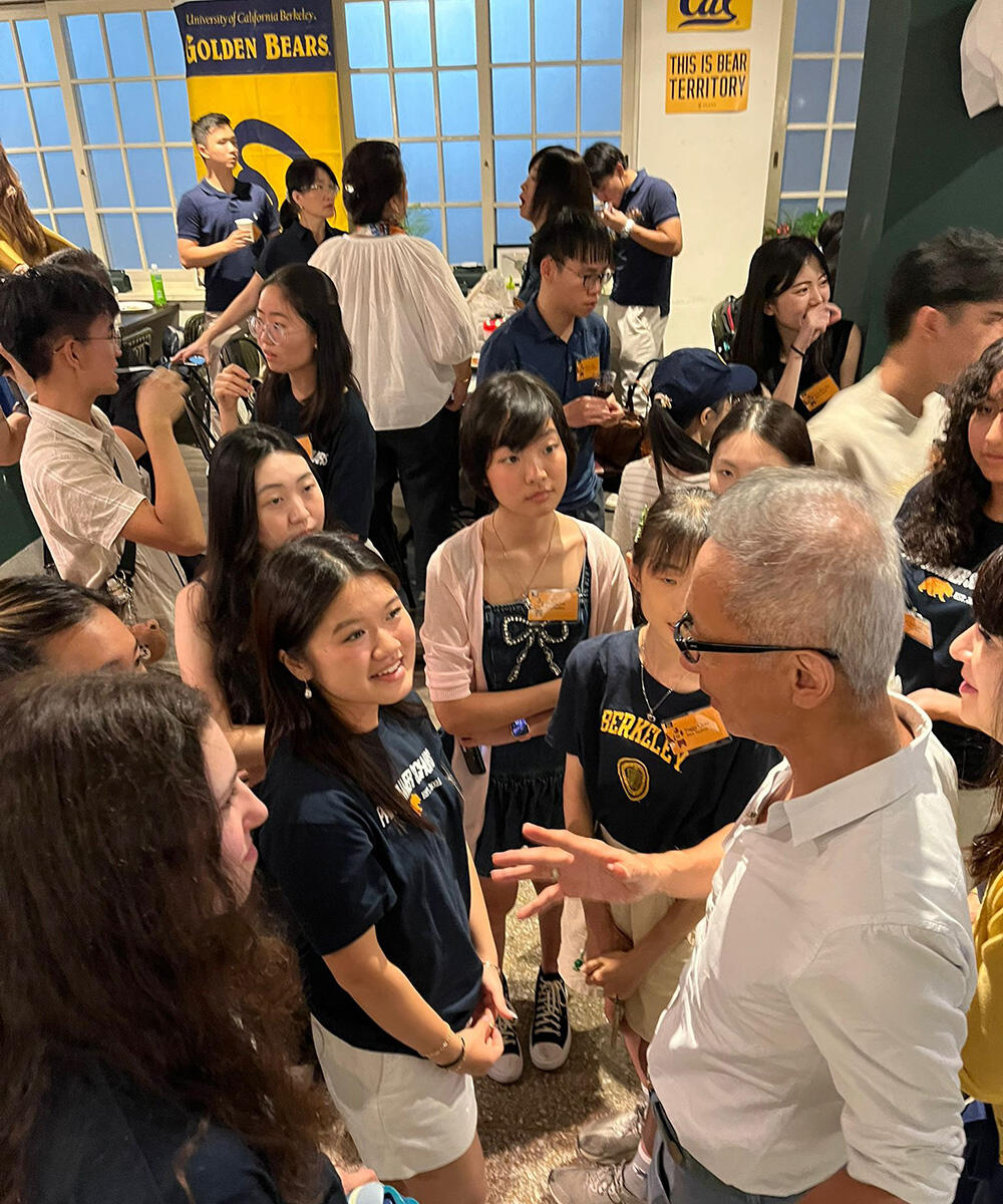 A group of students listen to a man in a white collared shirt at an event with Cal signs on the wall