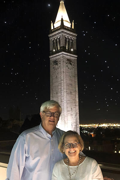 Two people stand on a rooftop balcony at night with a lit-up Campanile tower behind them