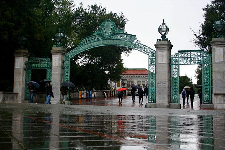 Rainy campus with Sather Gate in view, students walk around holding umbrellas