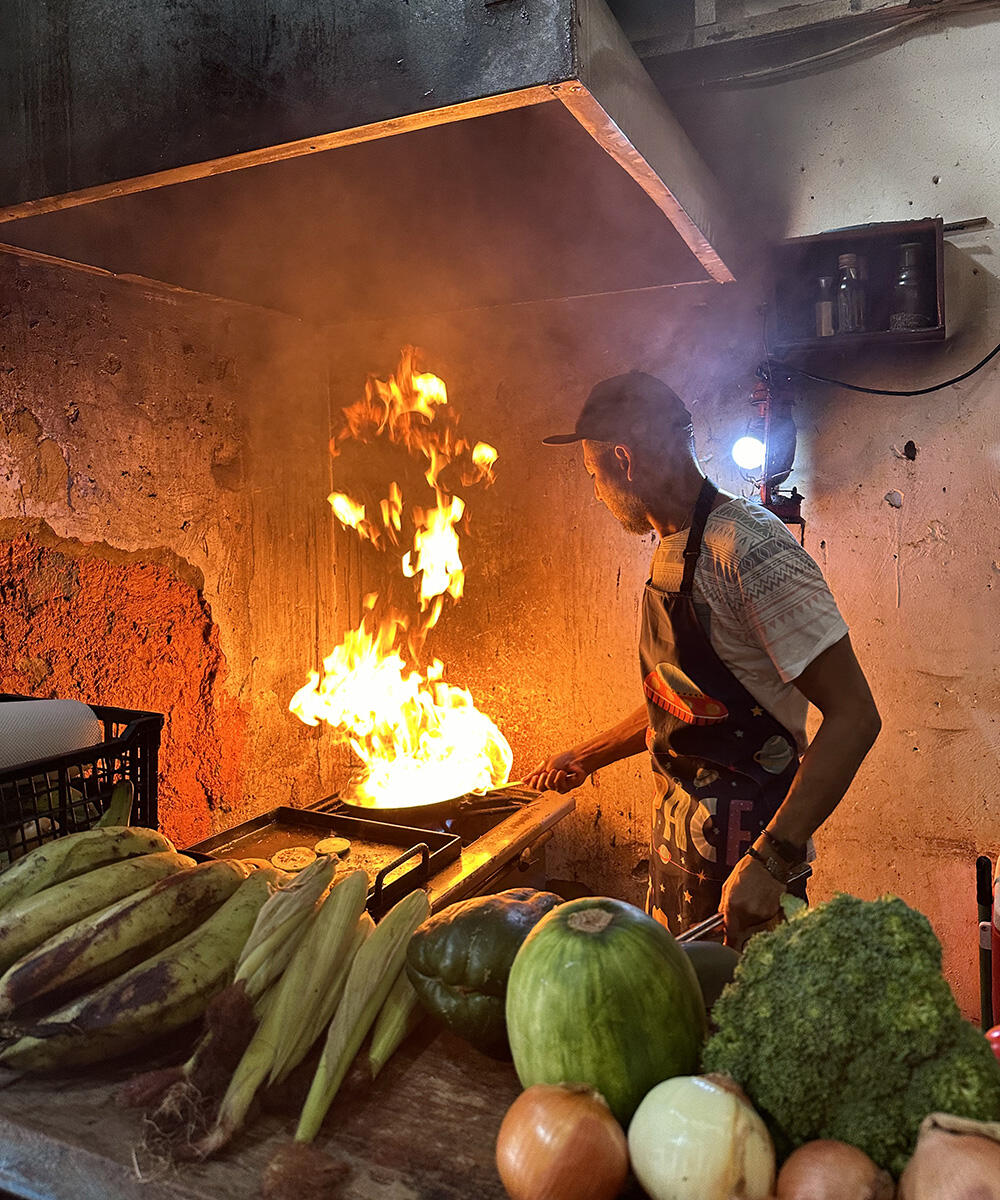 Tall flames erupt from a skillet as an apron-clad cook looks on. Produce sits on a shelf in the foreground.