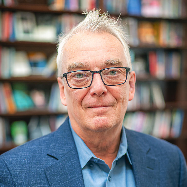 A headshot of a man in glasses and a suit jacket in front of full bookshelves