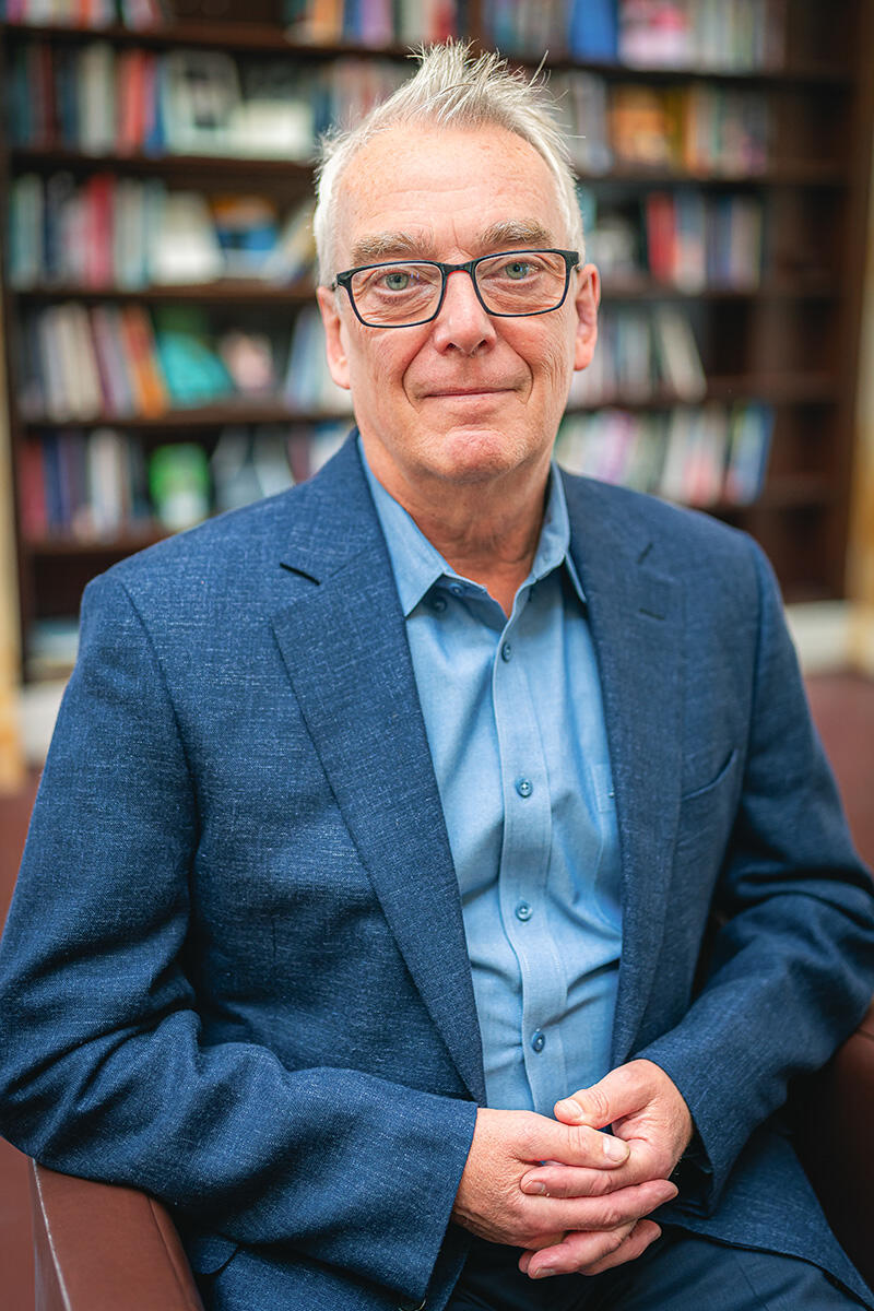 A man wearing a blue suit jacket and button-down shirt sits in front of a bookshelf