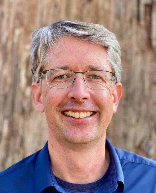 A man wearing a blue, buttoned shirt and glasses smiles toward the camera