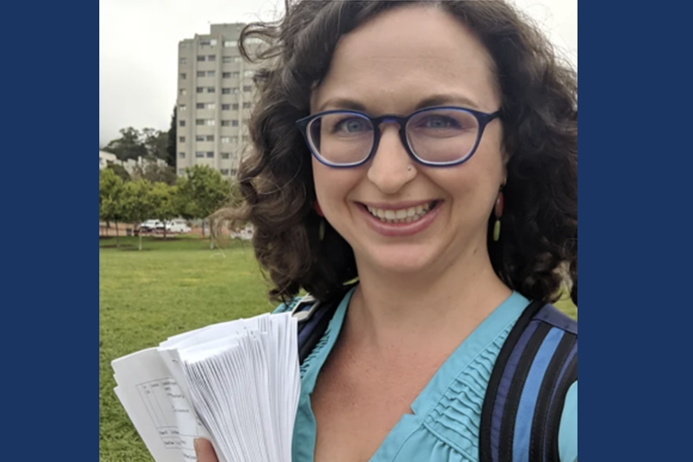 Sarah Gold McBride stands outside, brown curly hair and glasses, holding paper in hand.