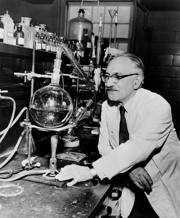 A black-and-white photo of a scientist in a lab coat looking at lab equipment
