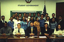 25 students in formal or semiformal attire pose behind a desk with the words "Student Government" on the wall behind them