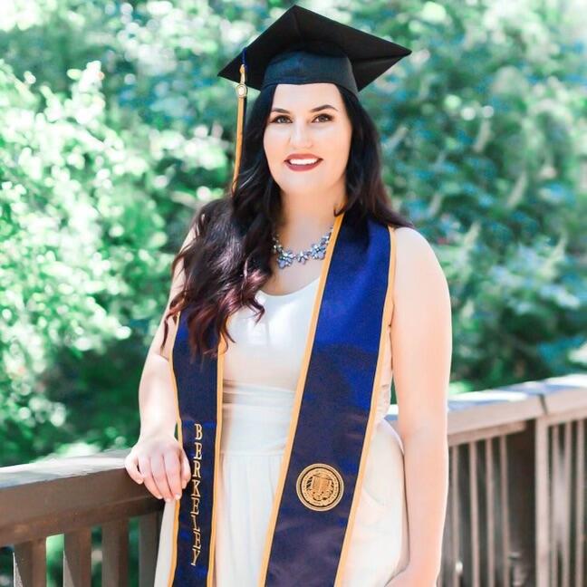 Woman with long dark hair wearing graduation cap and white dress, Berkeley regalia