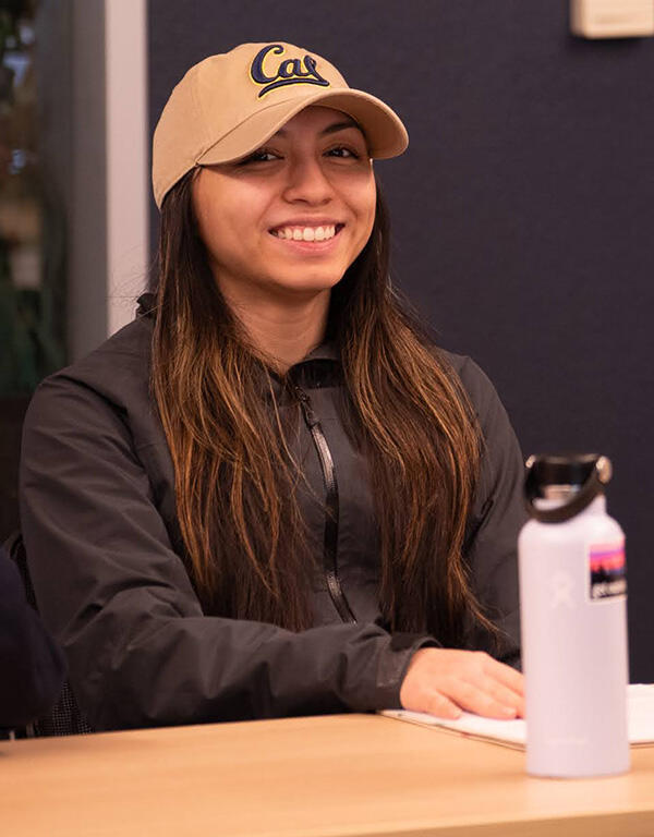 A woman in a Cal hat and dark jacket smiles at a table during a workshop.