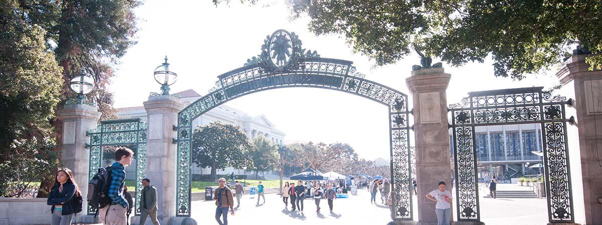 The sun hits Sather Gate as students walk by. In the foreground there is a male student with a backpack on the left side.