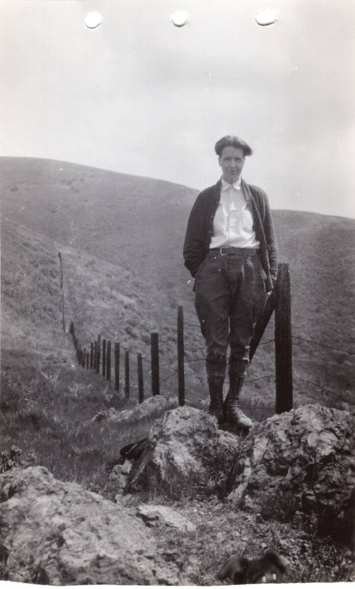 Black and white photo of woman with short hair wearing white button up shirt, cardigan, and trousers, standing on a rock near a fence outdoors