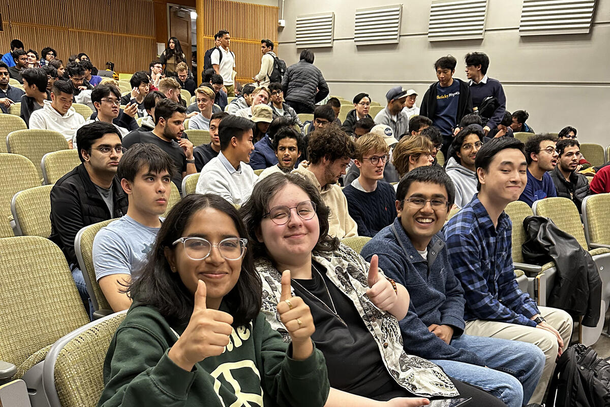 A crowd of students in an auditorium. Four are sitting in the front row smiling and raising their thumbs.