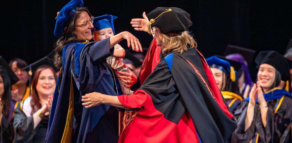 Graduate with child in arms embraces faculty member