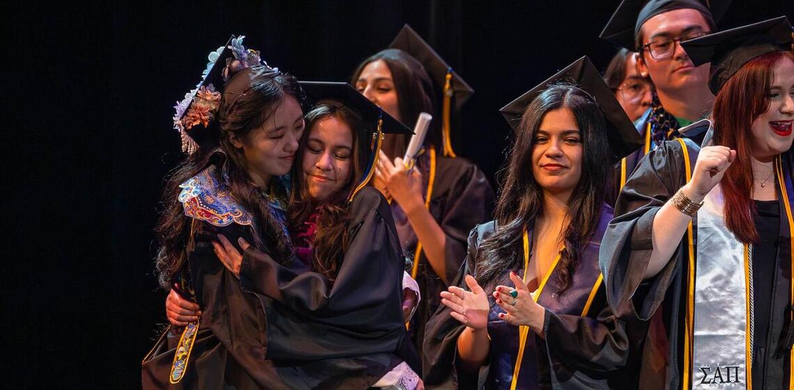 Graduates in regalia hug on stage