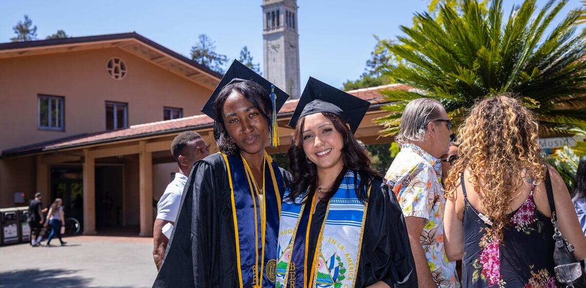 Two graduates pose for a photo with the Campanile in the background