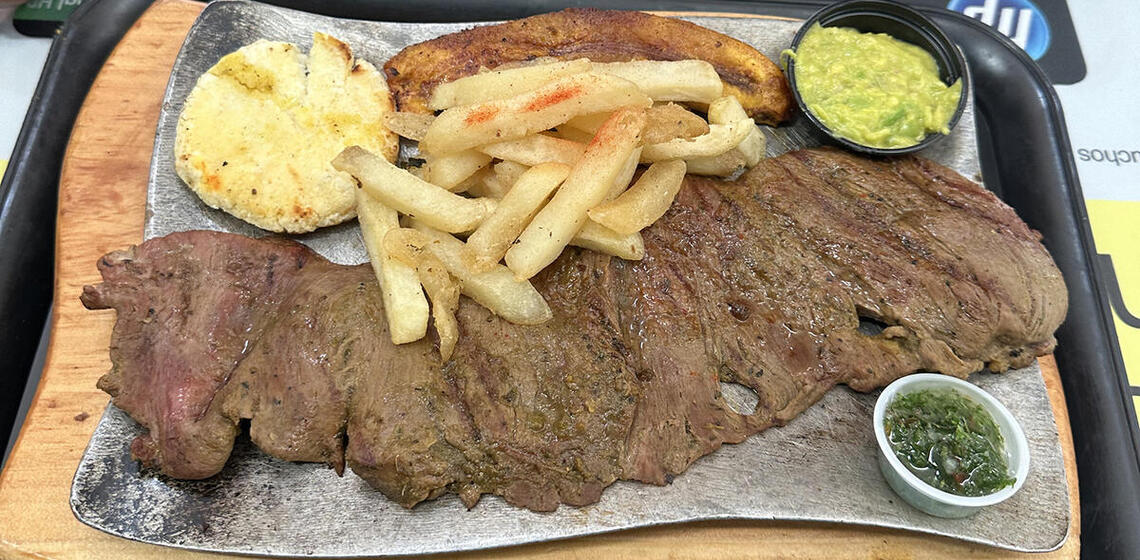 A large slab of meat with a plantain, fries, guacamole, and other items