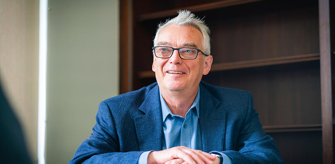 Man wearing blue shirt and suit jacket sits with arms folded at desk, in an office