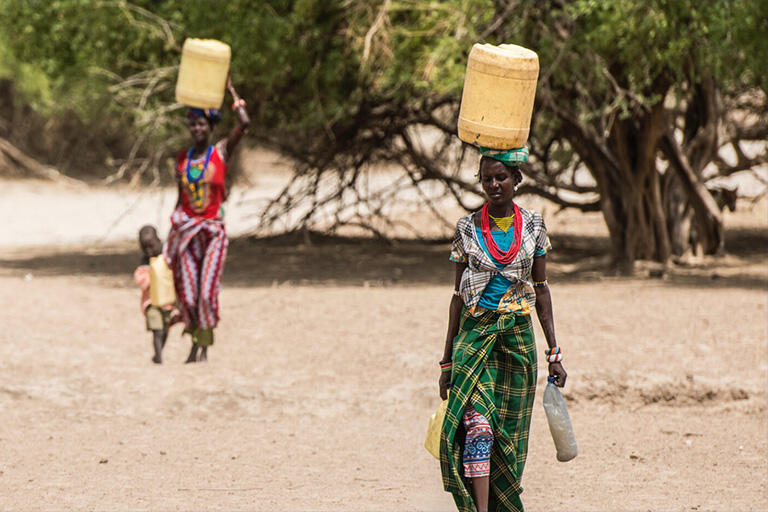 two women in colorful clothes walking through desert scrub, each carrying a plastic jerrycan on her head
