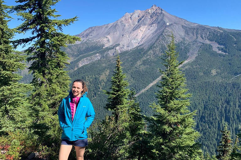 Person wearing shorts and a blue zip up jacket over a purple puffy vest, standing outdoors with a mountain peak behind them