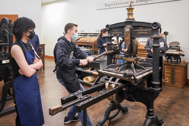 Student holds onto the lever of an old print press while other students watch. All students are wearing aprons and wearing face masks.
