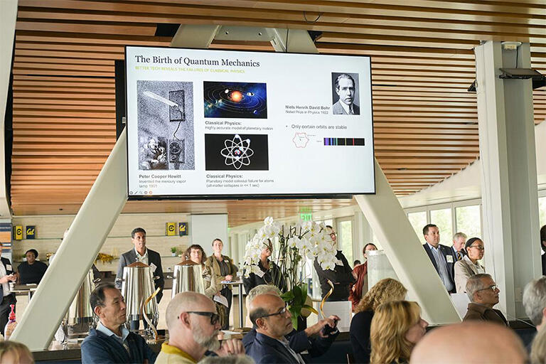 Group of people stand inside a conference room, with a large screen projecting "The Birth of Quantum Mechanics"