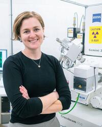 Headshot of Bronwyn Lucas with their arms crossed, standing in a lab in front of a machine