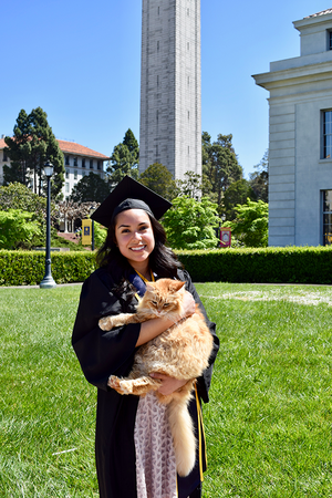 Graduate in gown poses in front of Campanile holding an orange cat