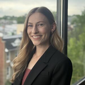 Headshot of Hannah Ousterman, person with long light brown hair wearing a dark blazer over a brown shirt