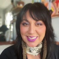 Headshot of a smiling person with long dark hair and bangs wearing a scarf and feather earrings