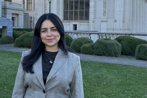 Woman wearing dark shirt under a black and white blazer stands outside in front of University library