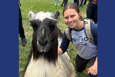 Woman wearing a backpack kneels on the ground, posing with a black and white llama