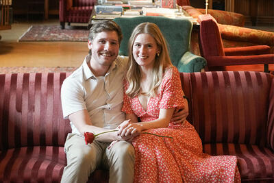 Couple sits on a couch, holding hands and a red rose, smiling