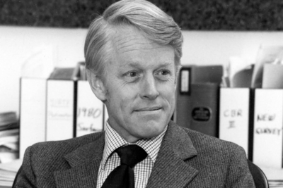 Black and white image of man wearing a tie, sitting in his office