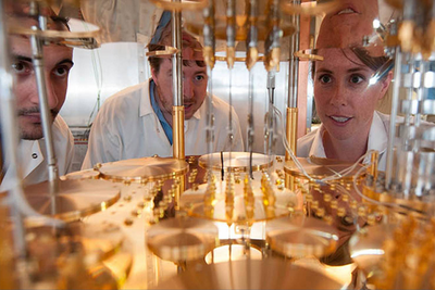 Three people in white lab coats peer into a very large, delicate, gold-colored machine.