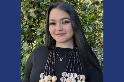 Headshot of person with long dark hair wearing a nose ring and strings of shells around their neck, along with a black shirt and silver necklace