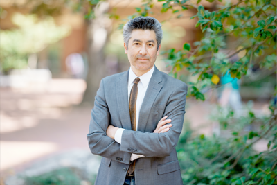 Man with dark hair stands outside with his arms crossed