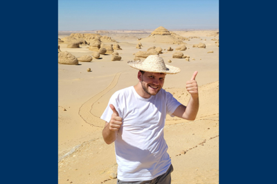 Man wearing straw hat and white tshirt gives two thumbs up while standing in desert