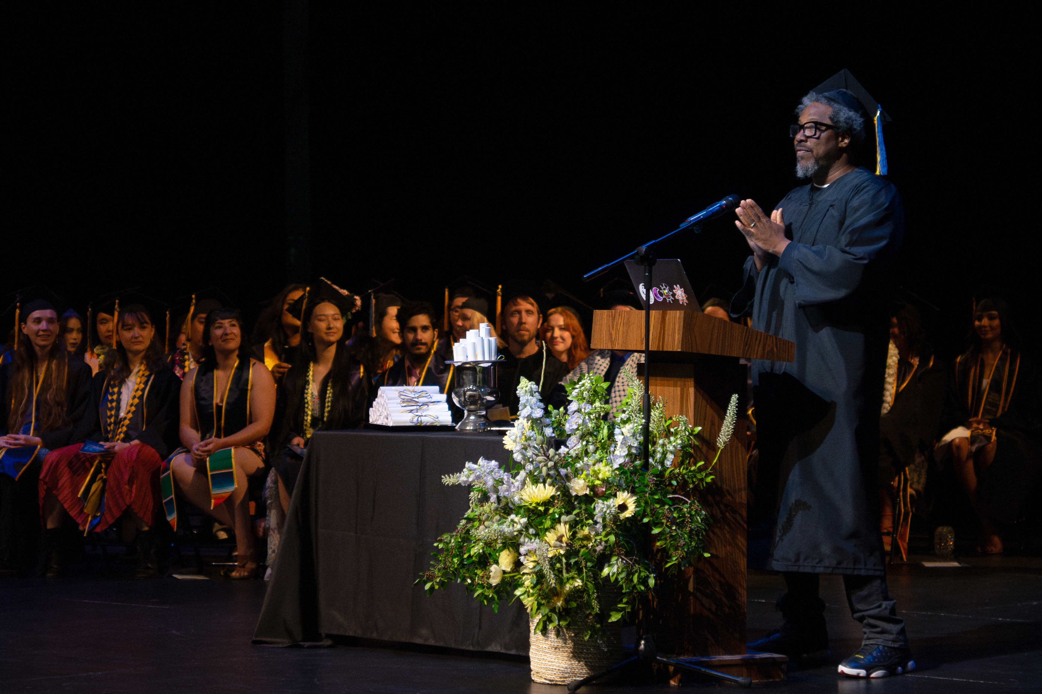 W Kamau Bell stands a podium during commencement, hands together, as graduates look on
